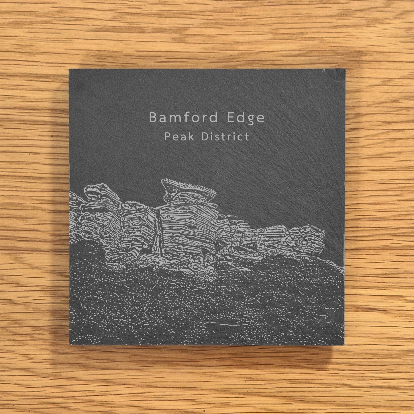Slate coaster with engraved image of Bamford Edge in the Peak District on a wooden surface