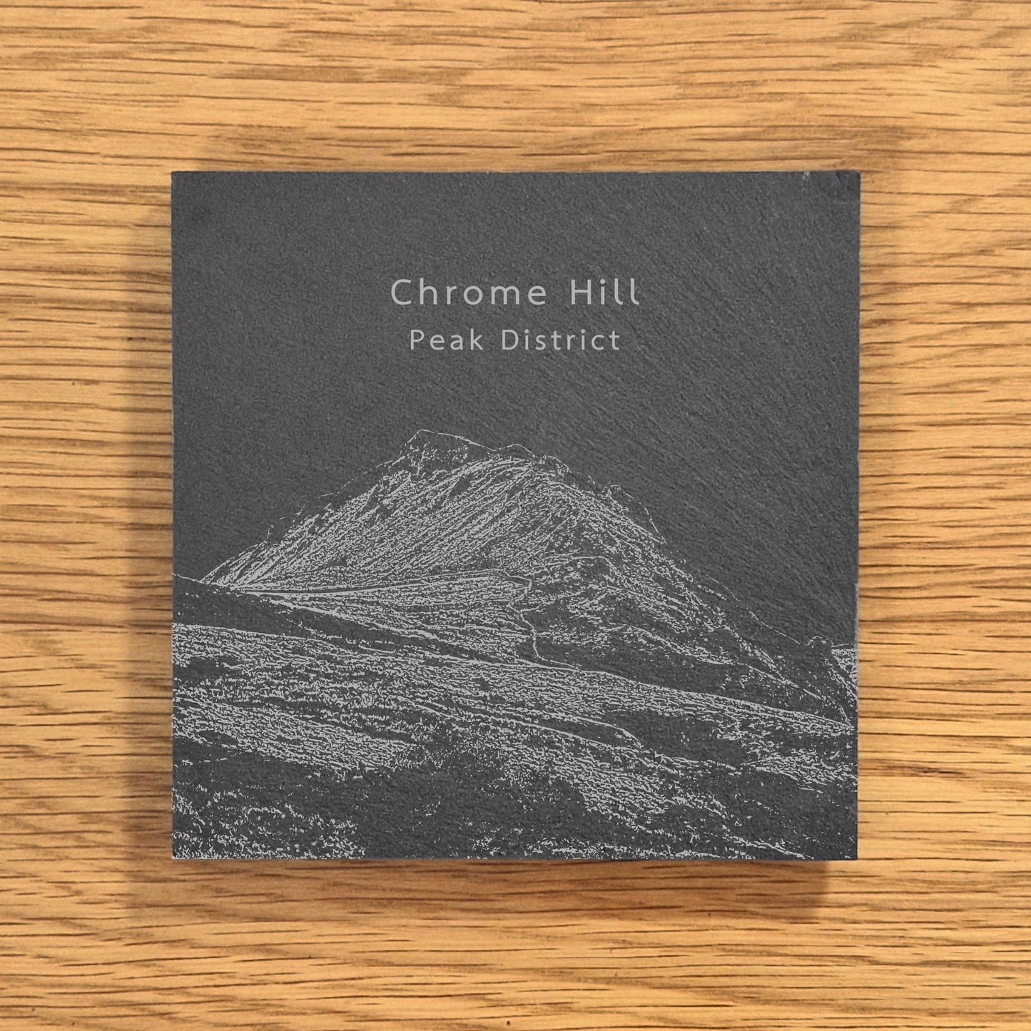 Slate coaster with engraved image of Chrome Hill (Dragon's Back) in the Peak District on a wooden surface.