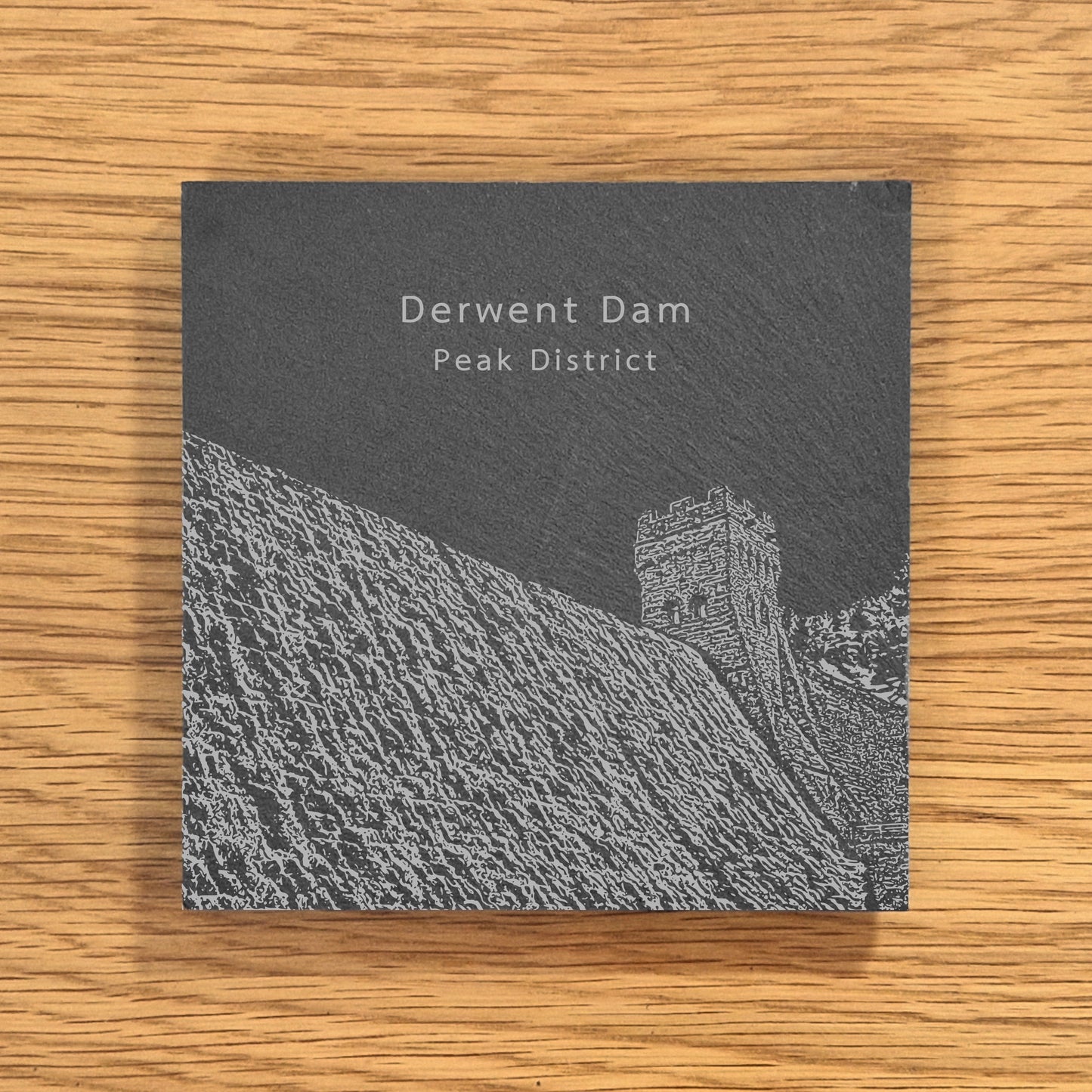 Slate coaster with an engraved design of Derwent Dam in the Peak District on a wooden surface
