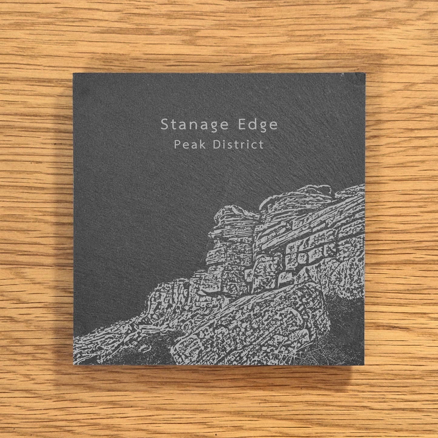 Slate coaster with engraved image of Stanage Edge in the Peak District on a wooden surface.