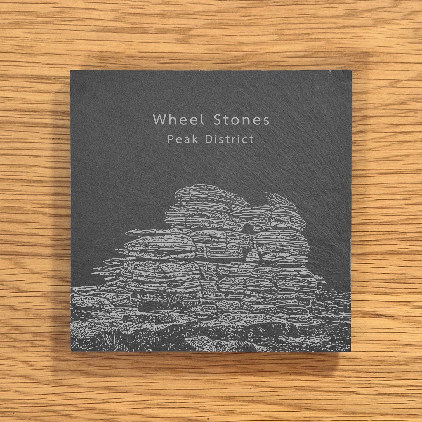 Slate coaster with engraved image of Wheel Stones in the Peak District on a wooden surface.