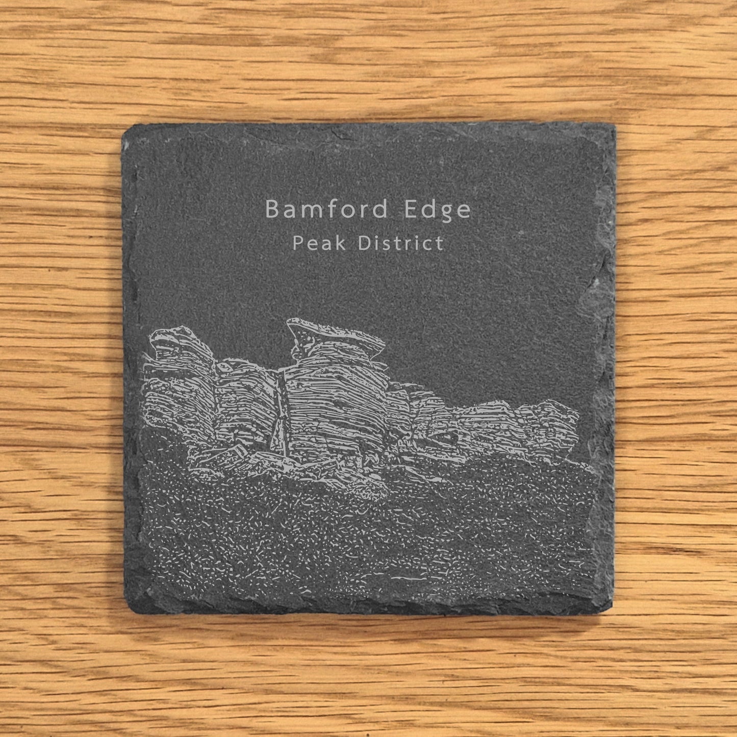 Slate coaster with engraved image of Bamford Edge in the Peak District on a wooden surface.