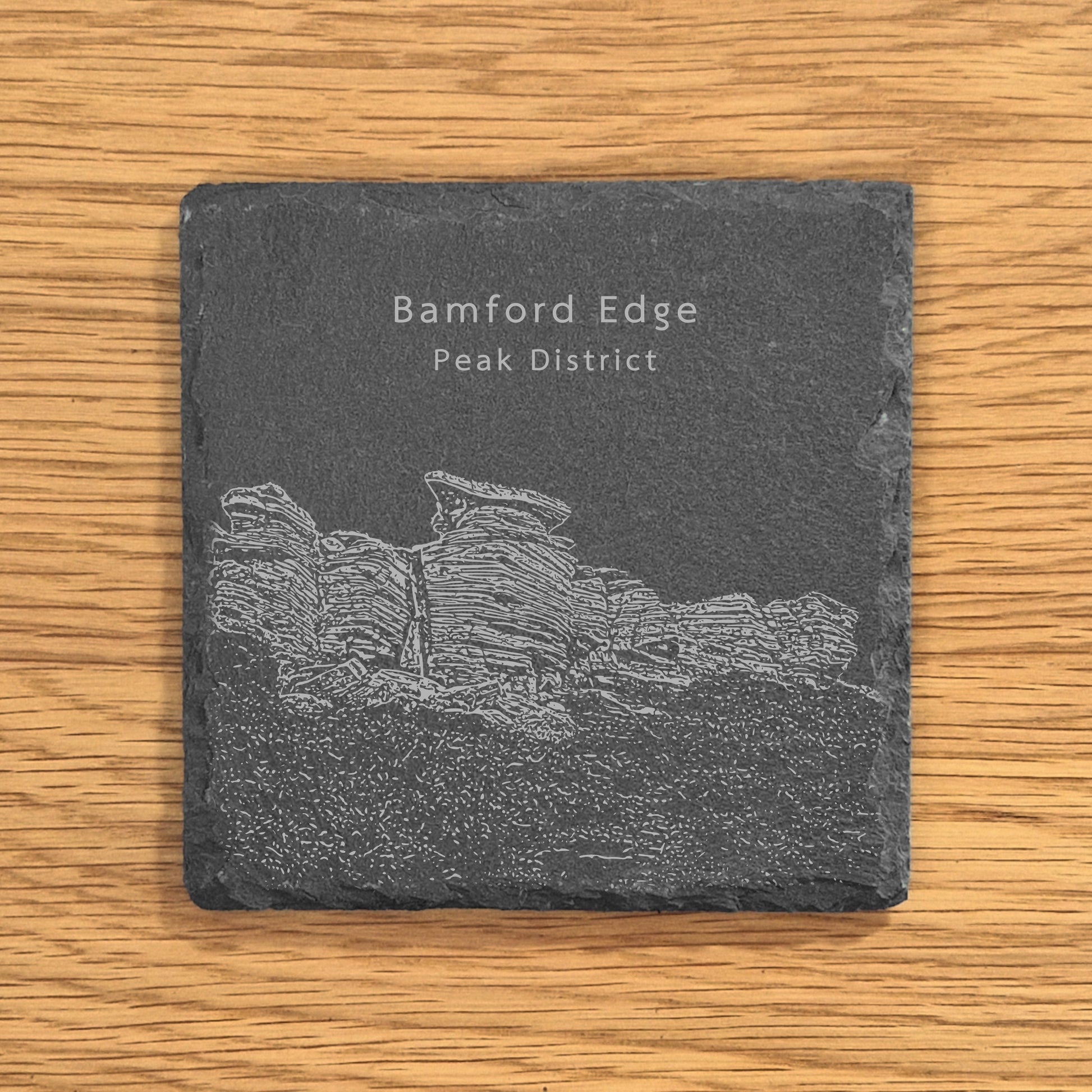 Slate coaster with engraved image of Bamford Edge in the Peak District on a wooden surface.