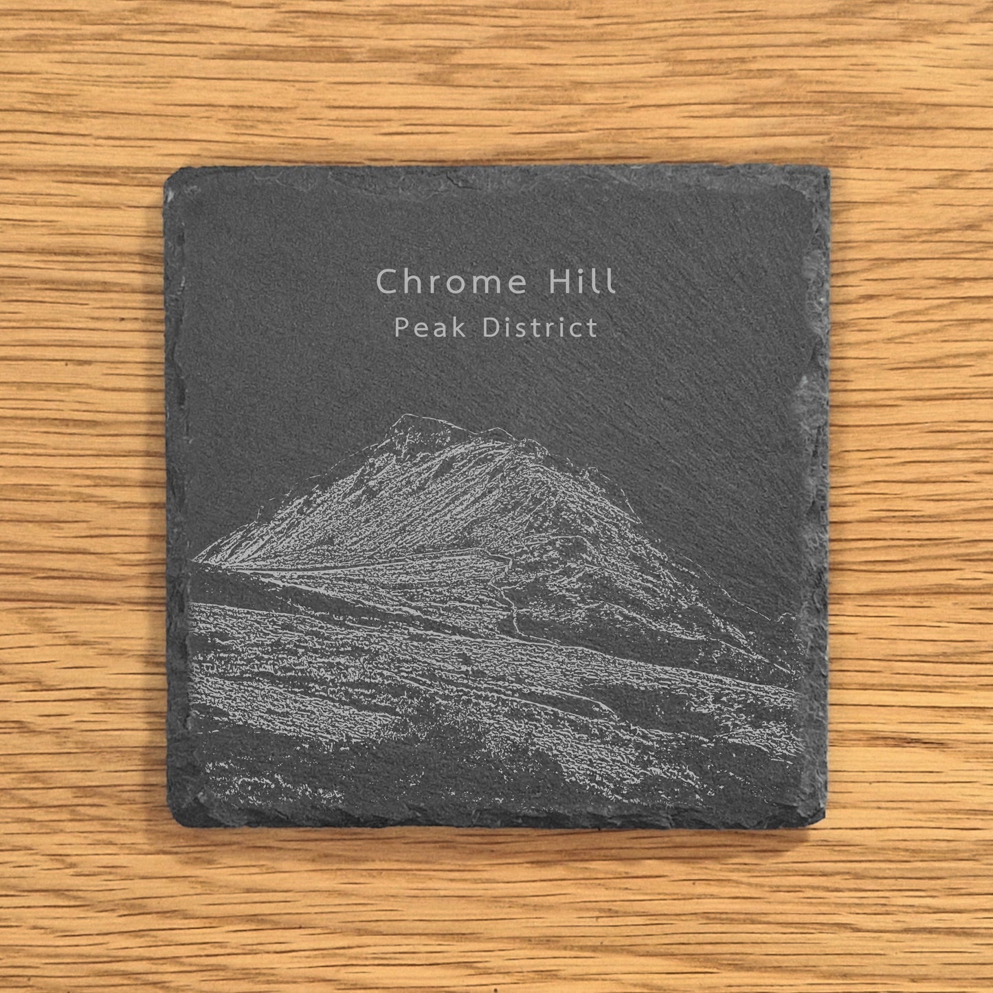 Slate coaster with engraved image of Chrome Hill (Dragon's Back) on a wooden surface.