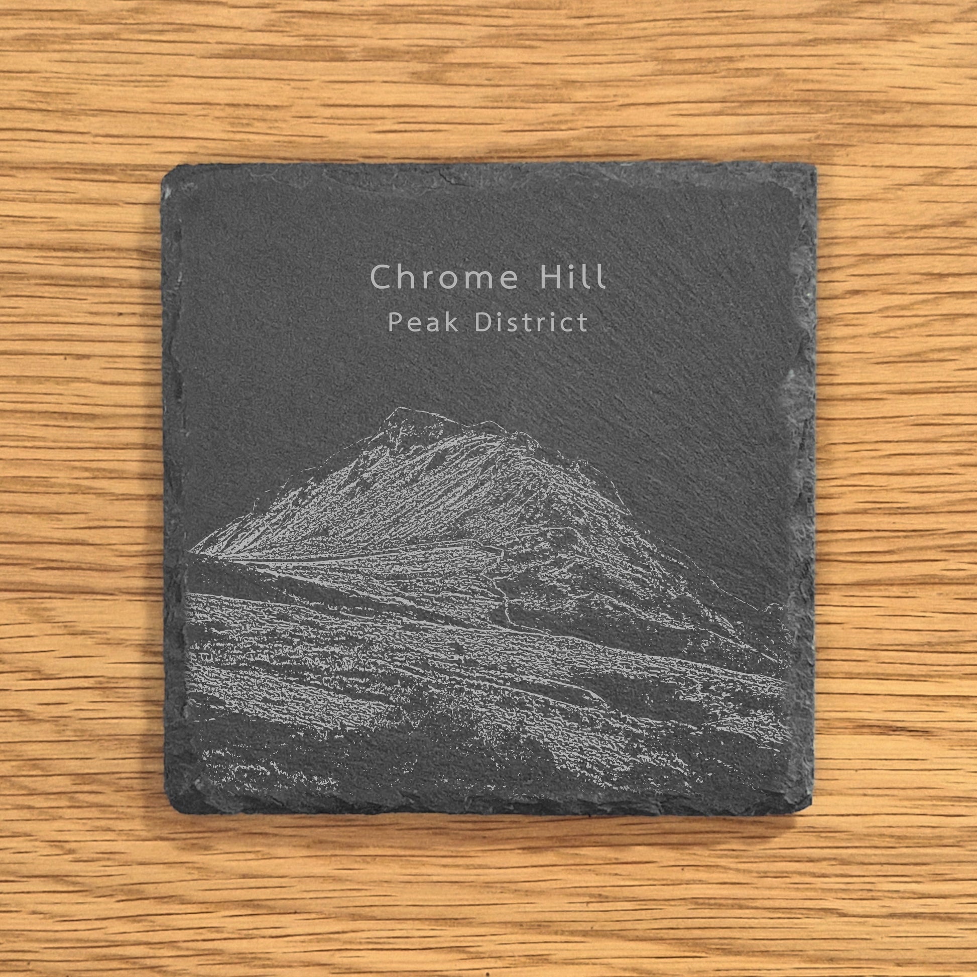 Slate coaster with engraved image of Chrome Hill (Dragon's Back) on a wooden surface.