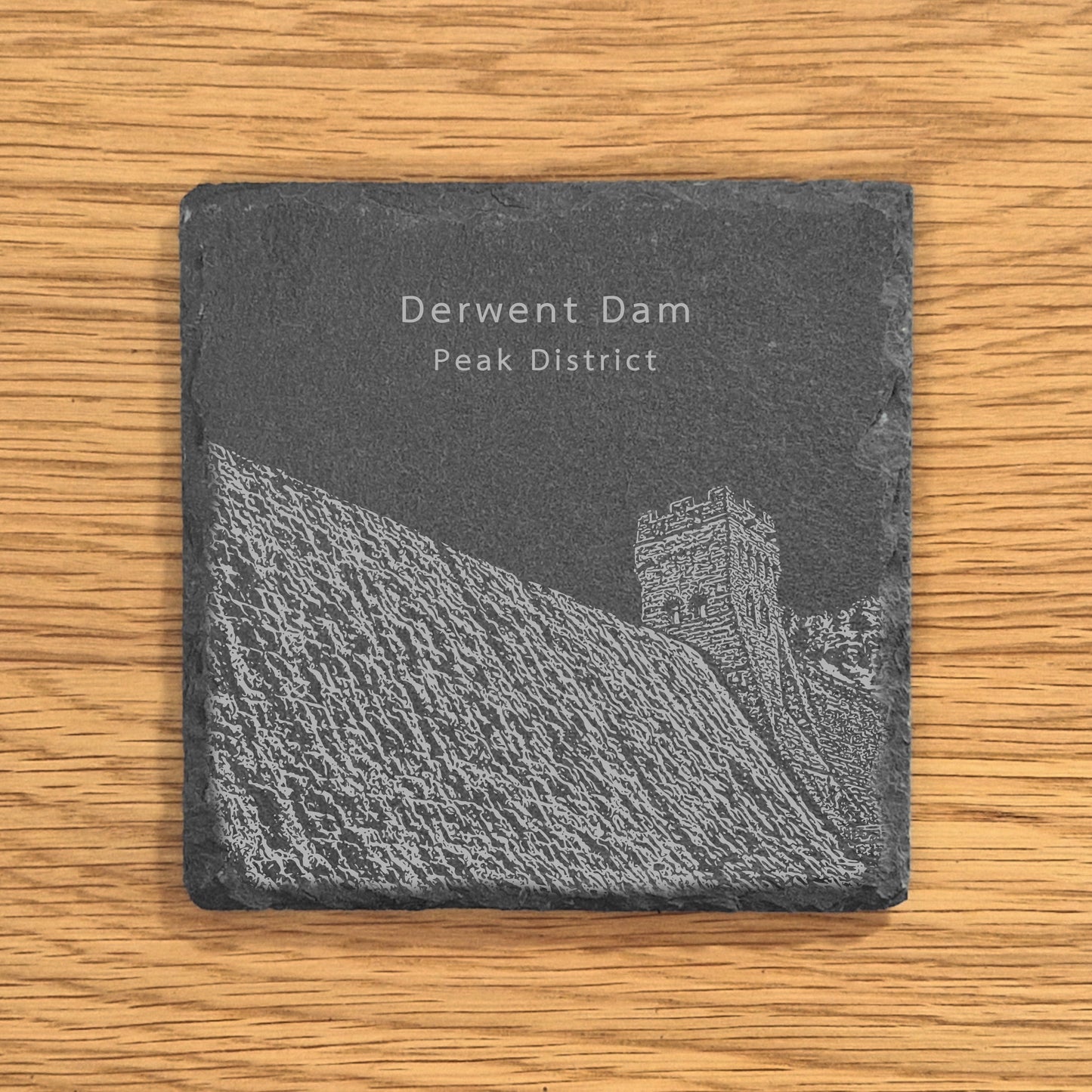 Slate coaster with an engraved design of Derwent Dam in the Peak District on a wooden surface.