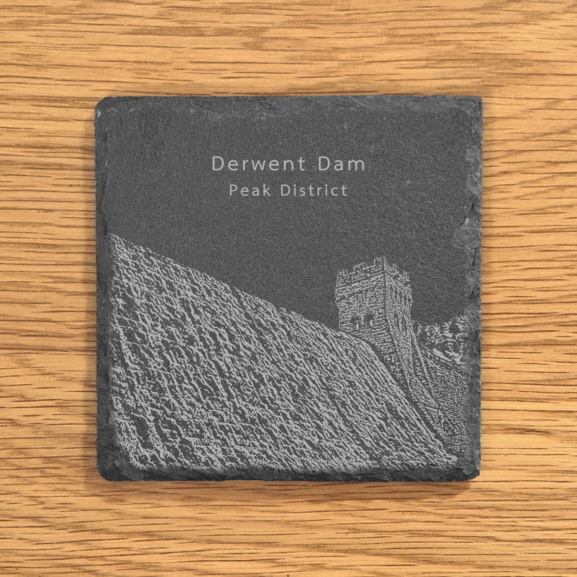 Slate coaster with an engraved design of Derwent Dam in the Peak District on a wooden surface.
