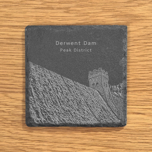 Slate coaster with an engraved design of Derwent Dam in the Peak District on a wooden surface.