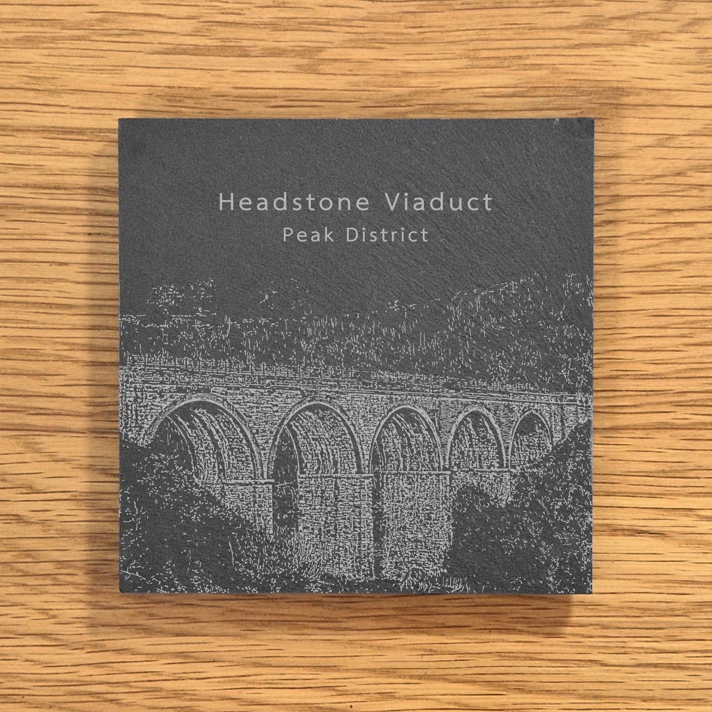 Slate coaster with engraved image of Headstone Viaduct in the Peak District on a wooden surface.