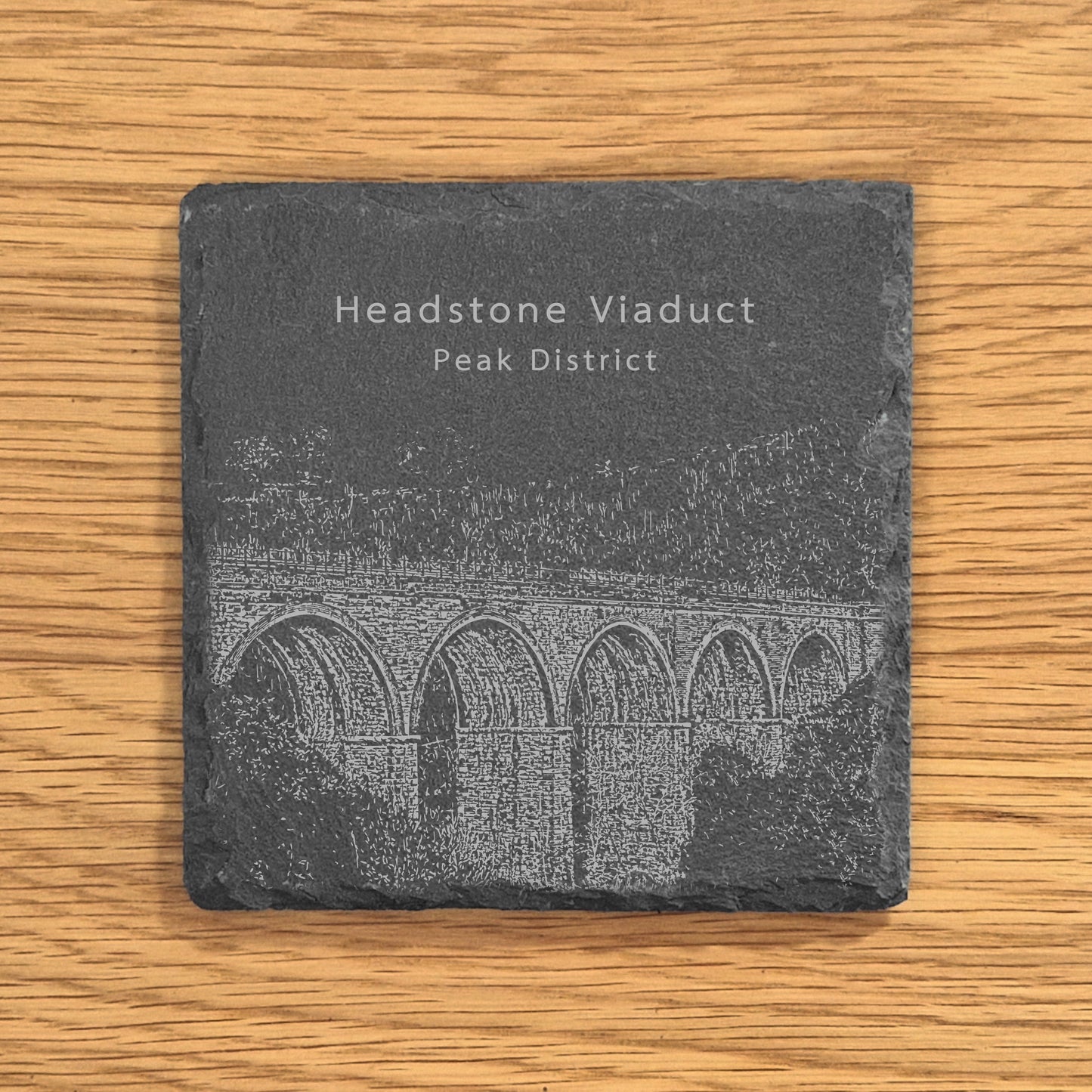 Slate coaster with engraved image of Headstone Viaduct in the Peak District on a wooden surface.