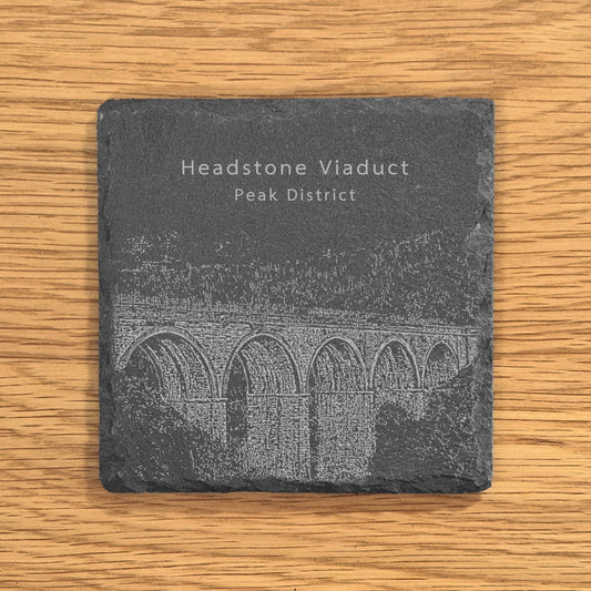 Slate coaster with engraved image of Headstone Viaduct in the Peak District on a wooden surface.