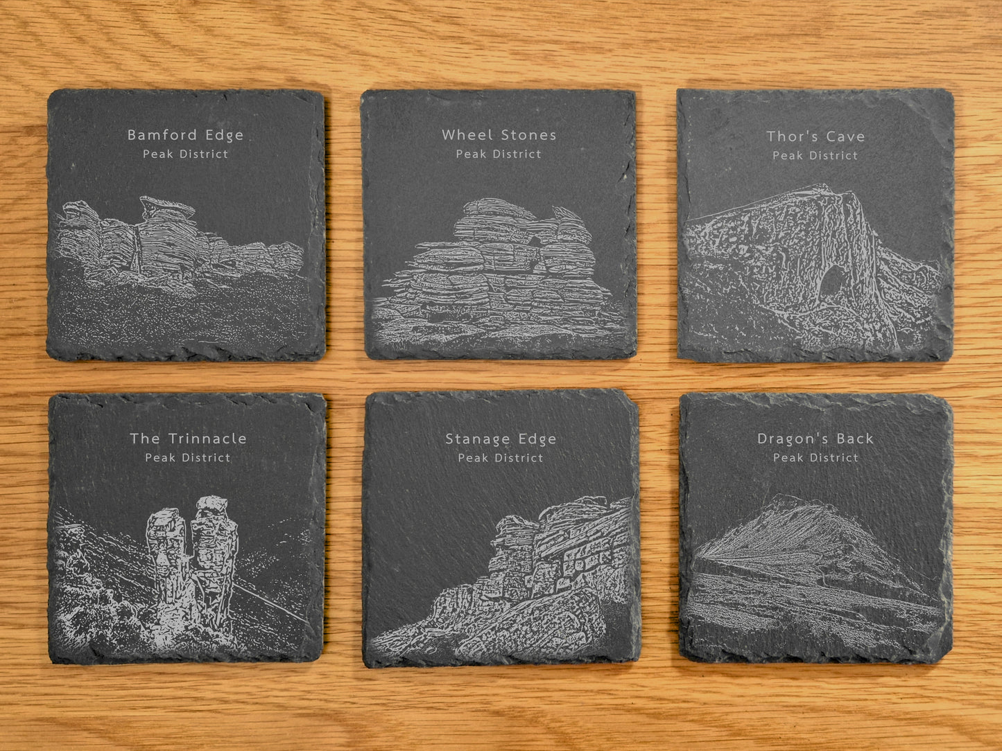 A set of six slate coasters, each engraved with a different Peak District landmark, displayed on a wooden surface.