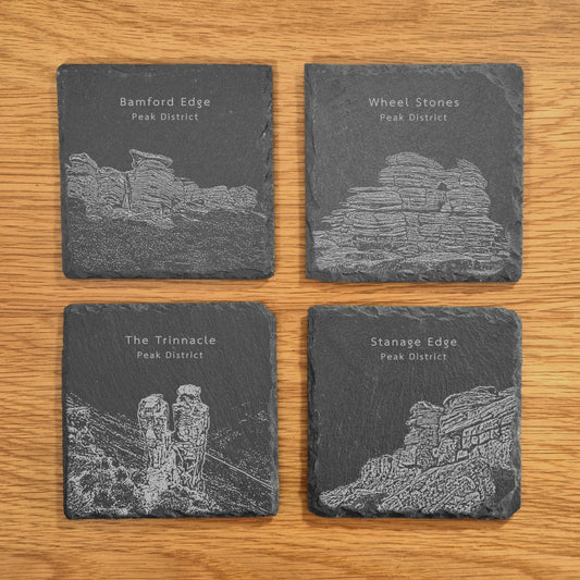 A set of four slate coasters, each engraved with a different Peak District landmark, Bamford Edge, Wheel Stones, The Trinnacle and Stanage Edge, displayed on a wooden surface.