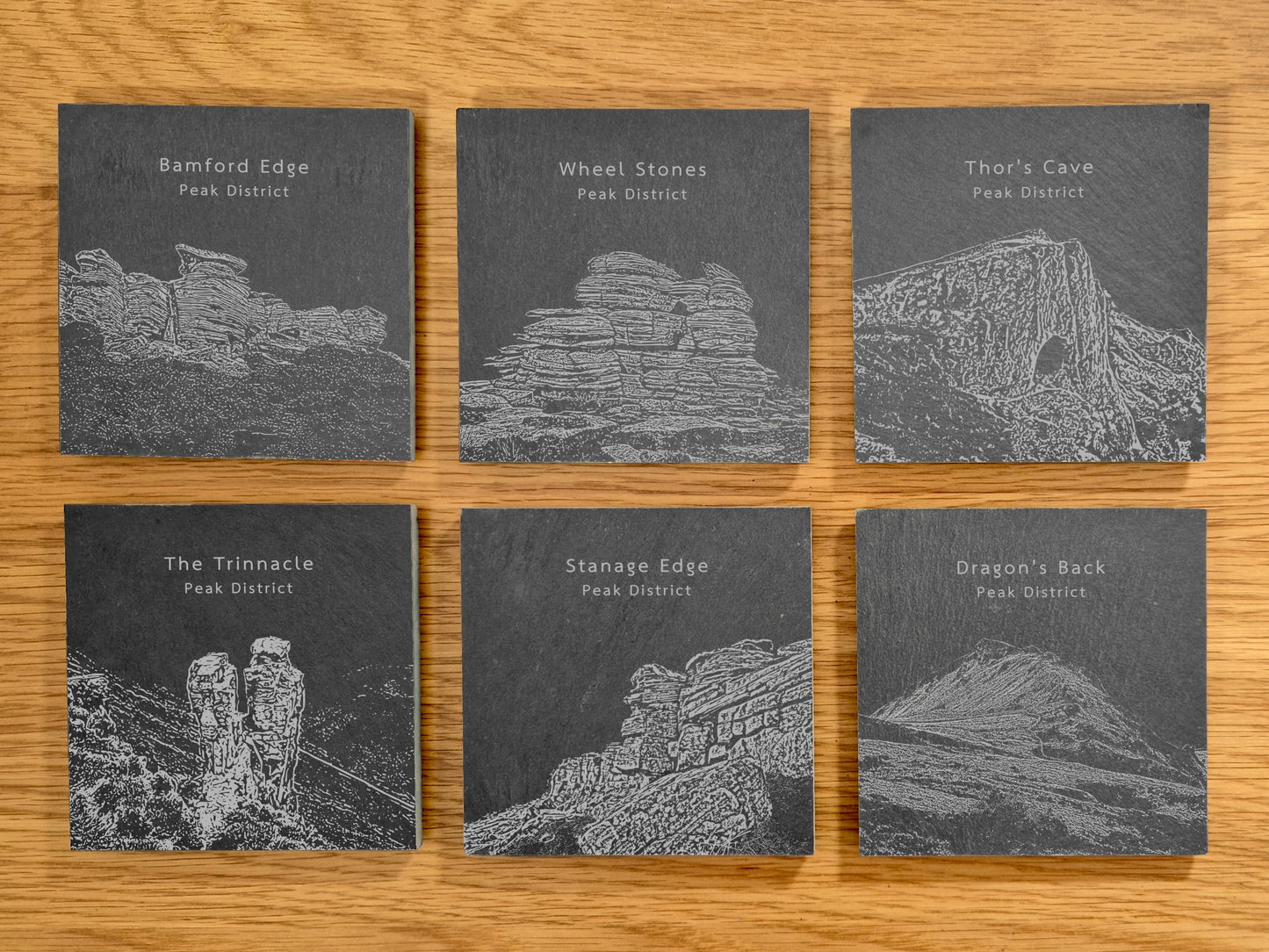 Six slate coasters engraved Peak District landmarks on a wooden surface