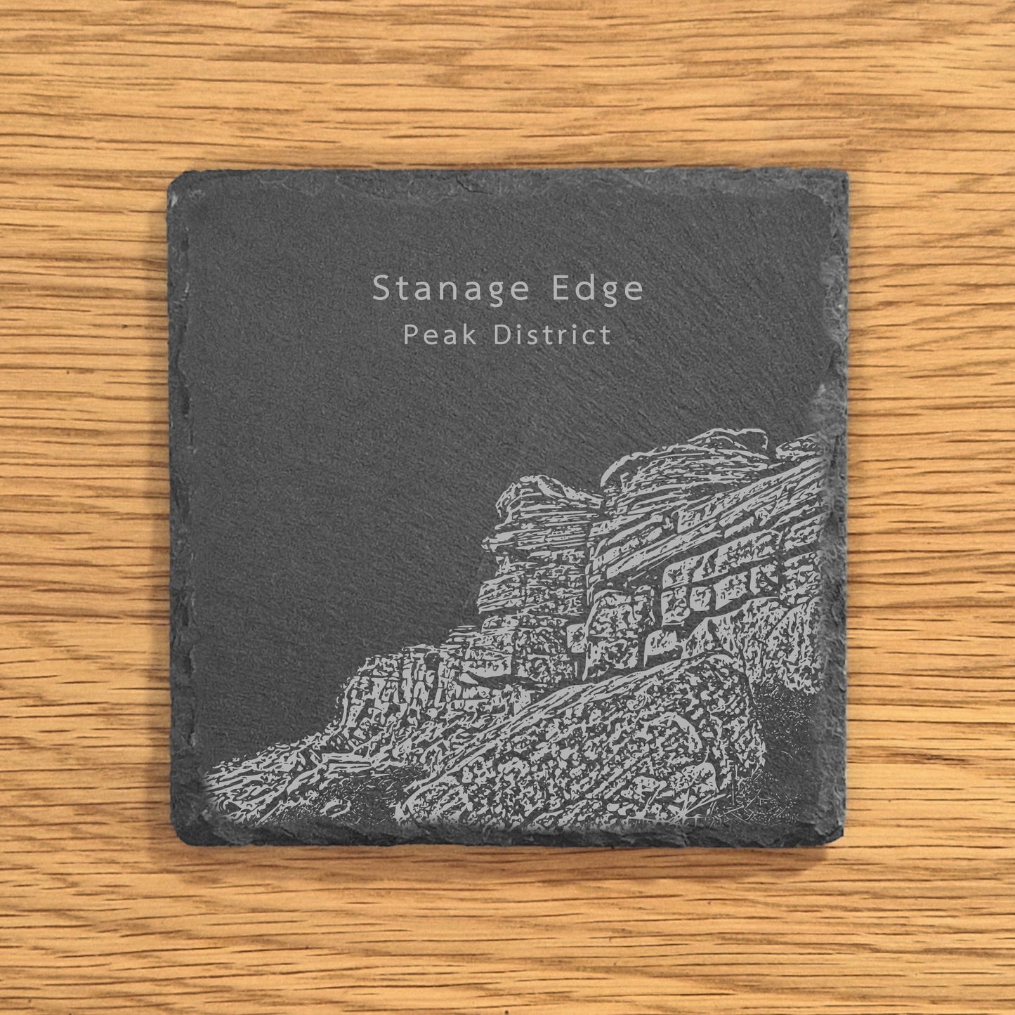 Slate coaster with engraved image of Stanage Edge in the Peak District on a wooden surface.