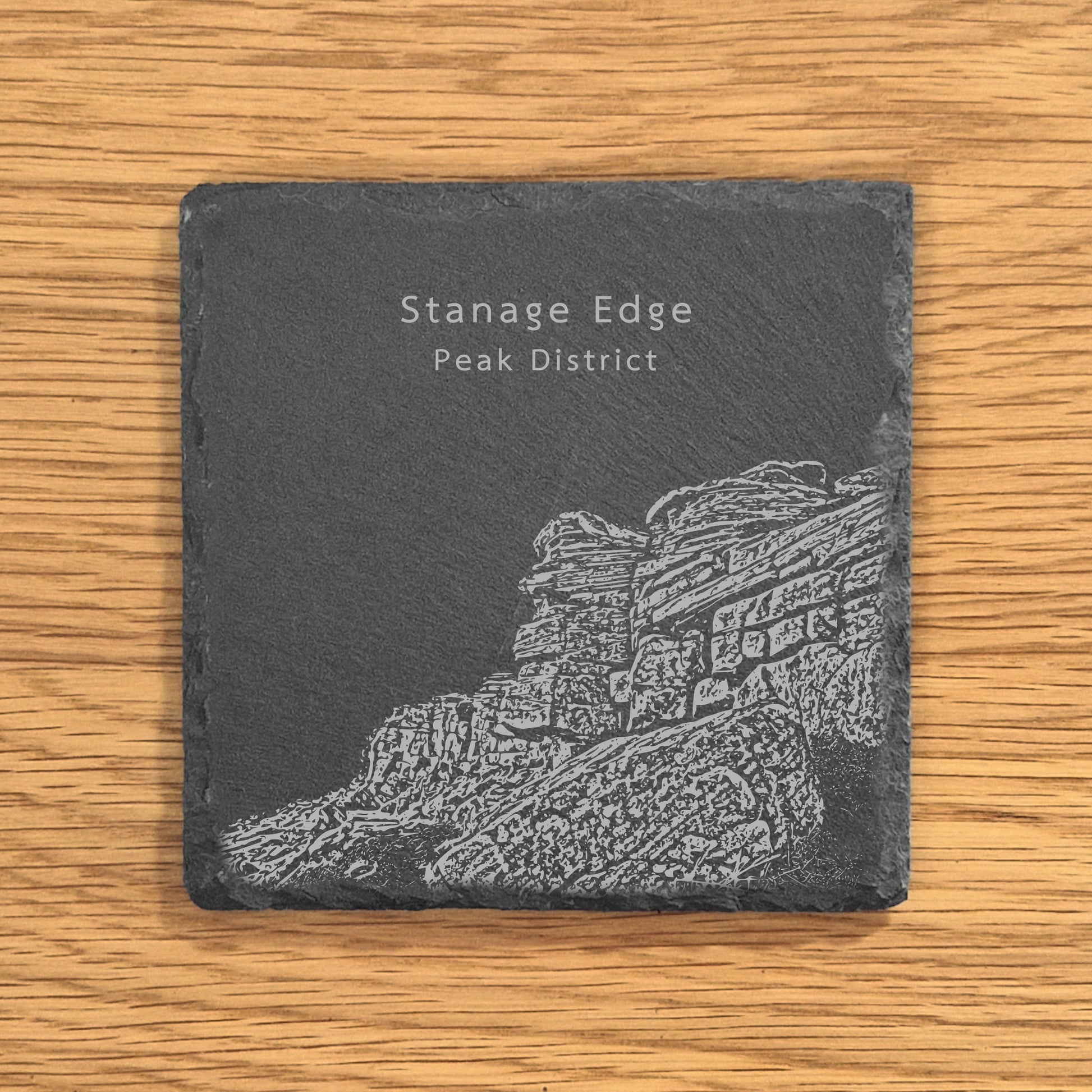 Slate coaster with engraved image of Stanage Edge in the Peak District on a wooden surface.