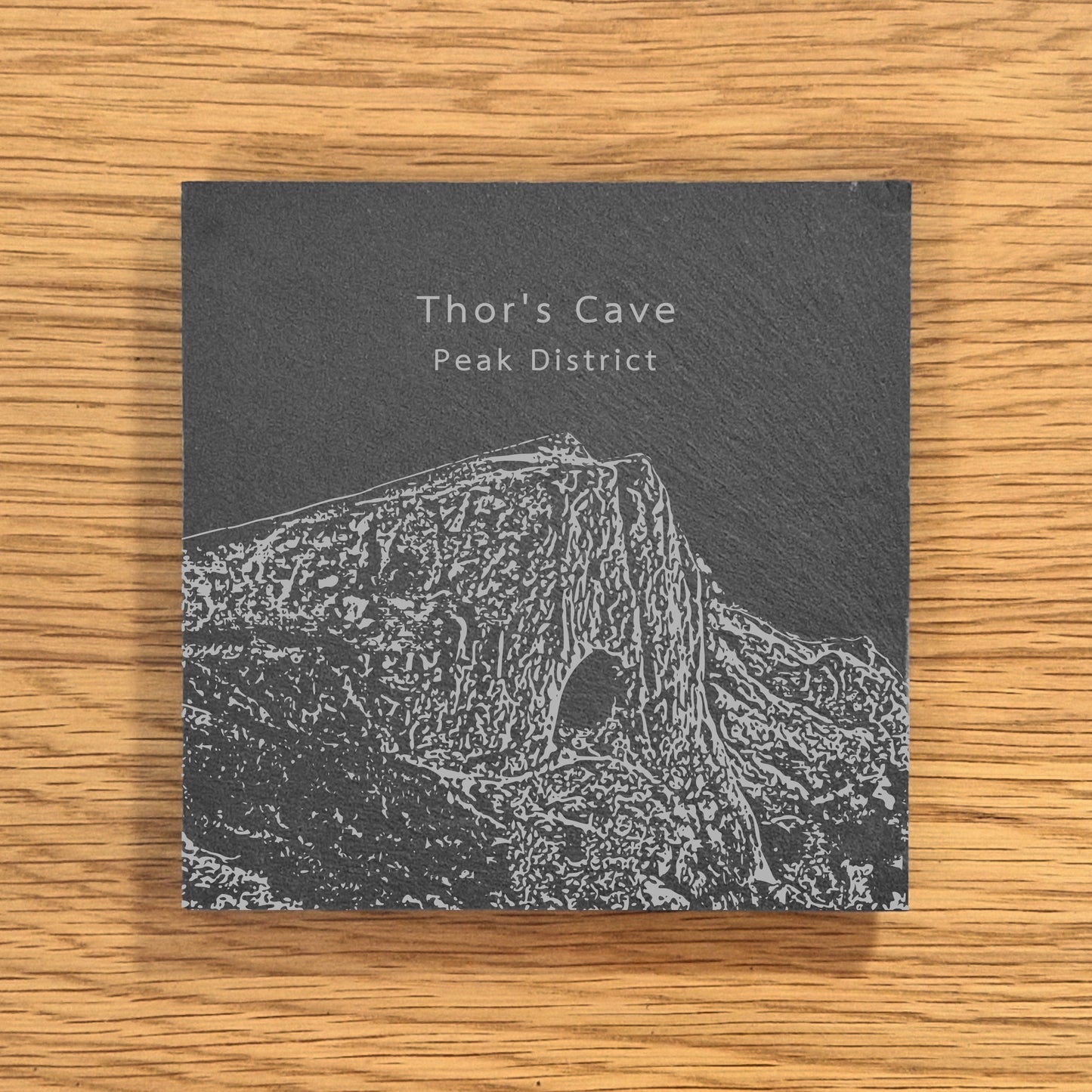 Slate coaster with engraved design of Thor's Cave in the Peak District on a wooden surface.
