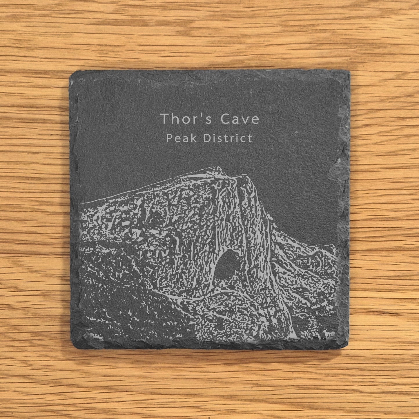 Slate coaster with engraved image of Thor's Cave in the Peak District on a wooden surface.