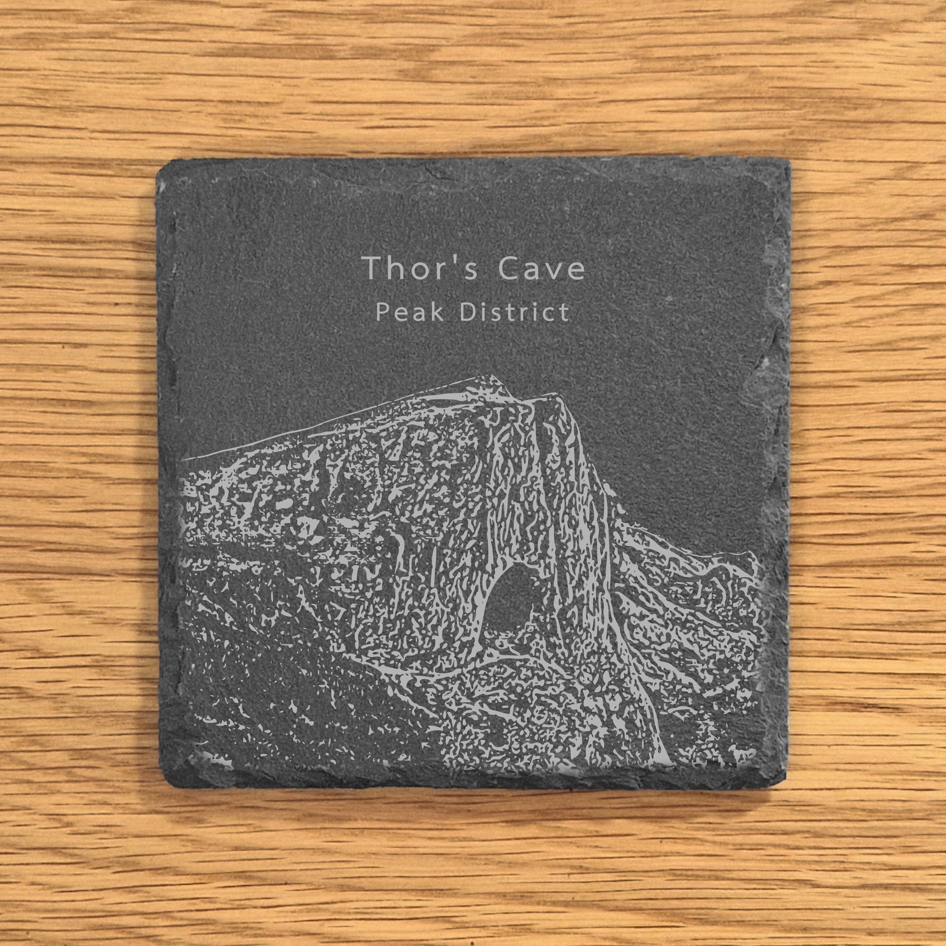 Slate coaster with engraved image of Thor's Cave in the Peak District on a wooden surface.