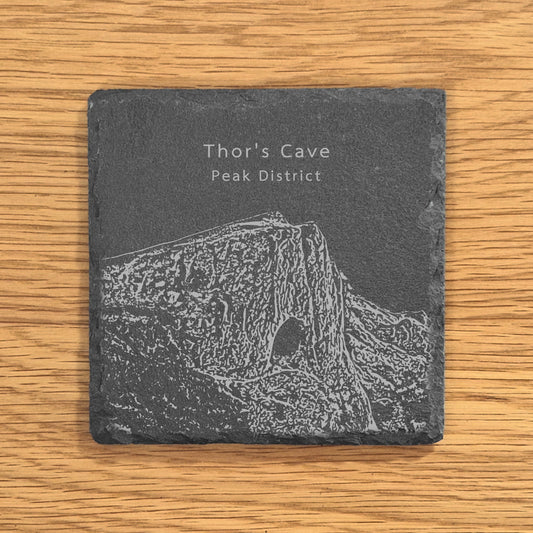 Slate coaster with engraved image of Thor's Cave in the Peak District on a wooden surface.